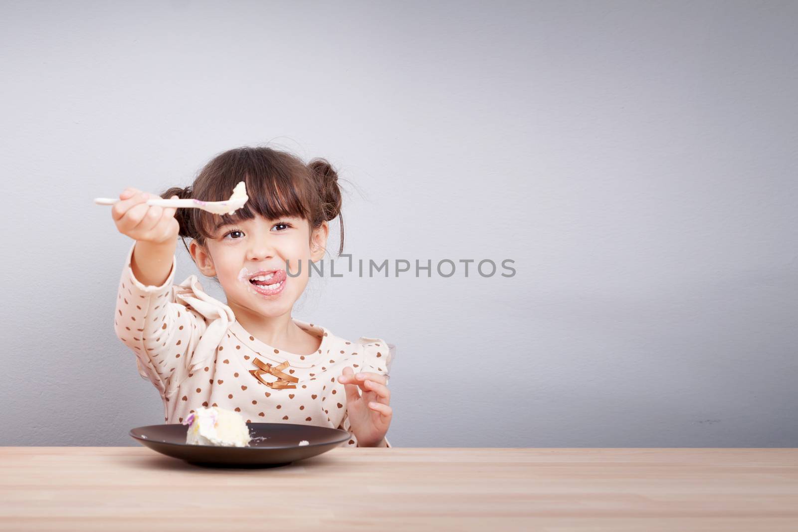 Kids enjoy eating concept : Happy little cute mixed race girl enjoy eating cake with smiley face , tongue stick with spoon in her hand for invite to eat. Kid poster background.