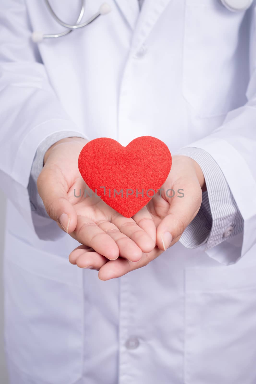 Young Asian doctor stand holding a red heart with two hands. Dress up with doctor gown uniform with stethoscope in a hospital. Beautiful Asia female model in her 20s , anonymous face