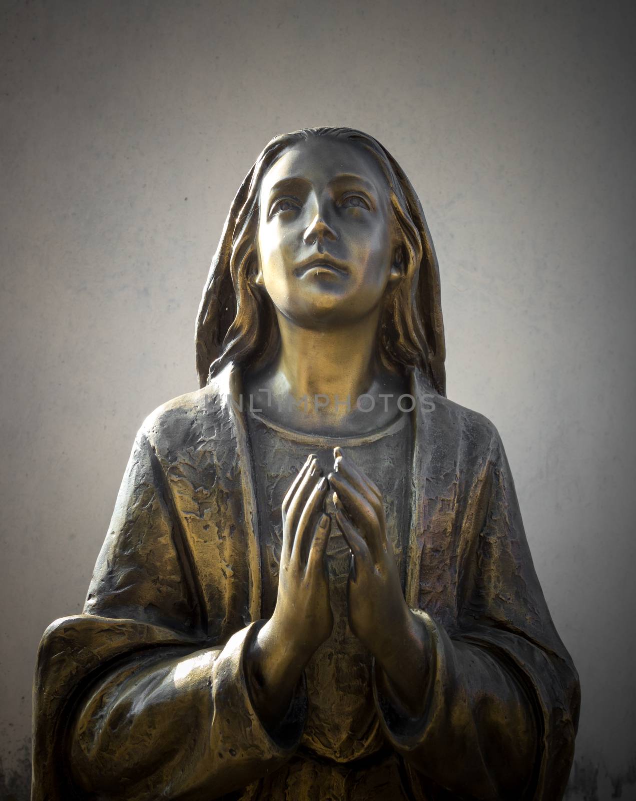 Holy Mary statue bronze that prays with folded hands, in the background a gray wall. It can be used for events, concepts and backgrounds.