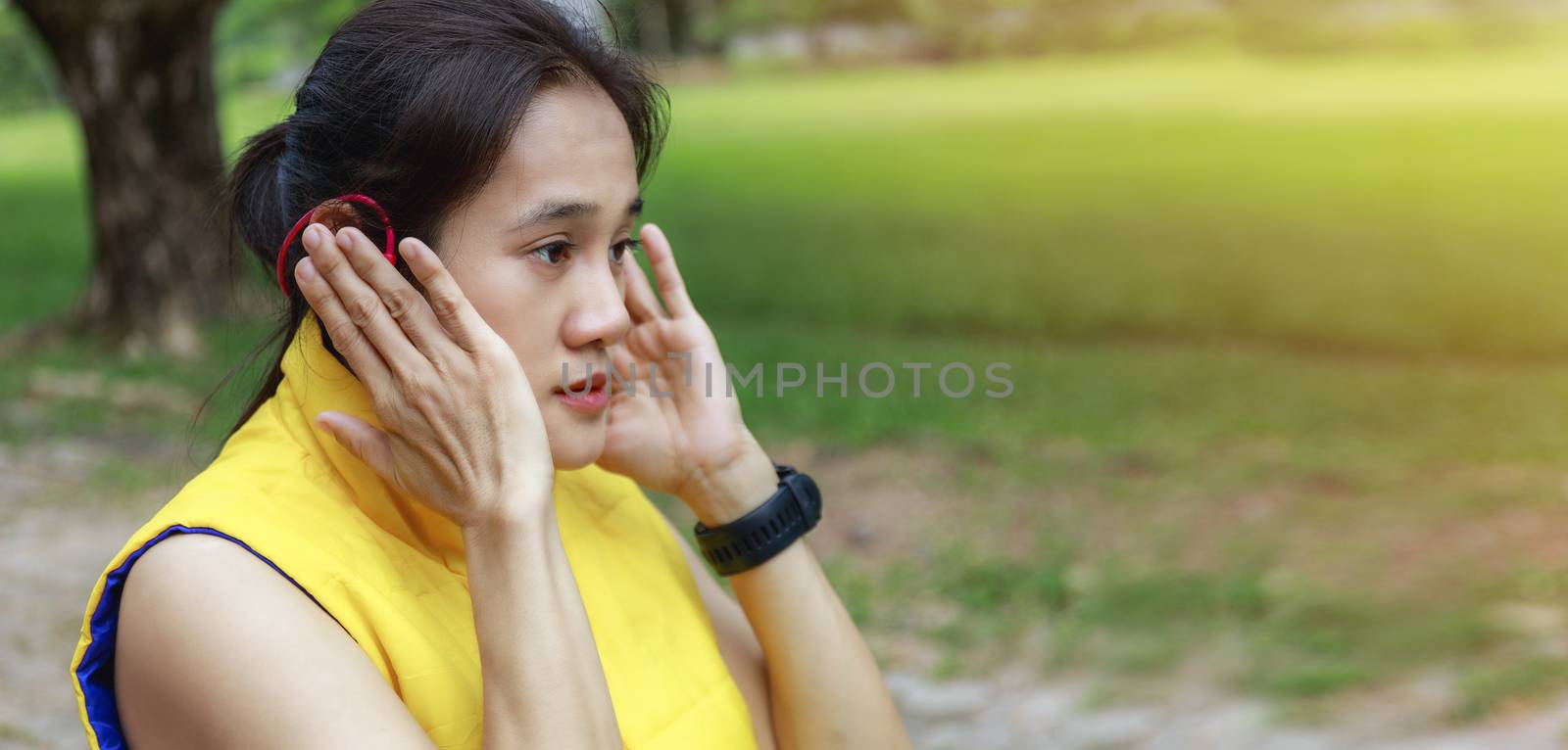 Asian woman long hair wearing yellow jacket relax  listening to music at park after running. Young woman feeling relaxed after long run during sunflair. Sport women relax with music after running with copy space banner. Women walk relax two hand hold music portable player in ear.