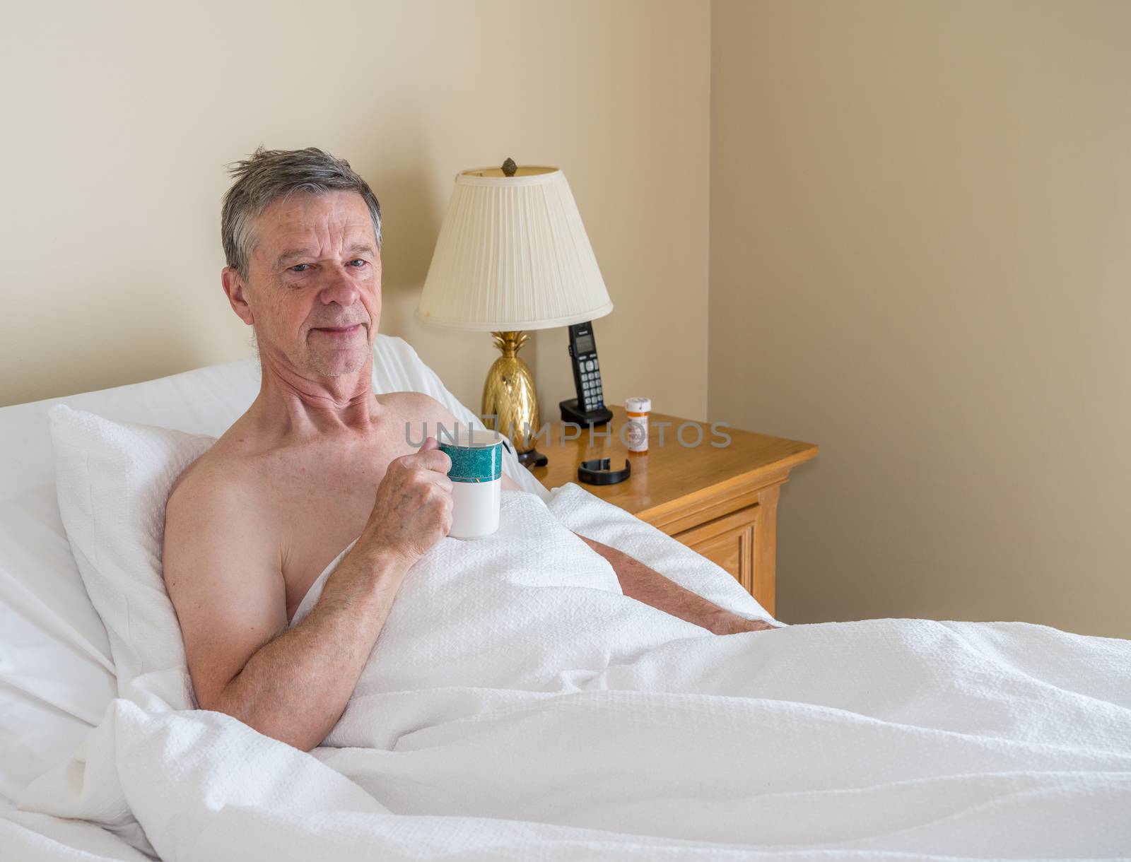 Senior retired caucasian man lying in adjustable bed on incline. He has a cup of coffee and is smiling towards the viewer