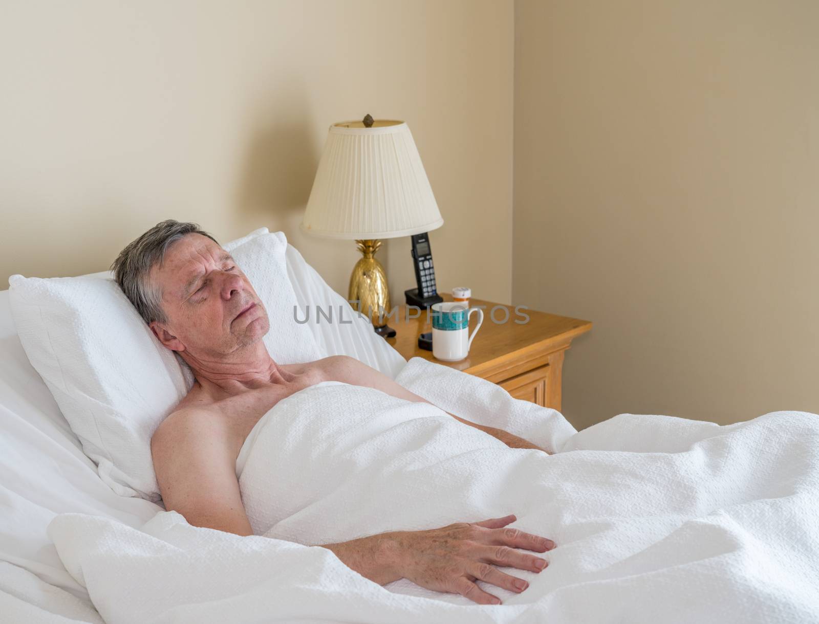Senior retired caucasian man lying in adjustable bed on incline. He is asleep with cup of coffee on table
