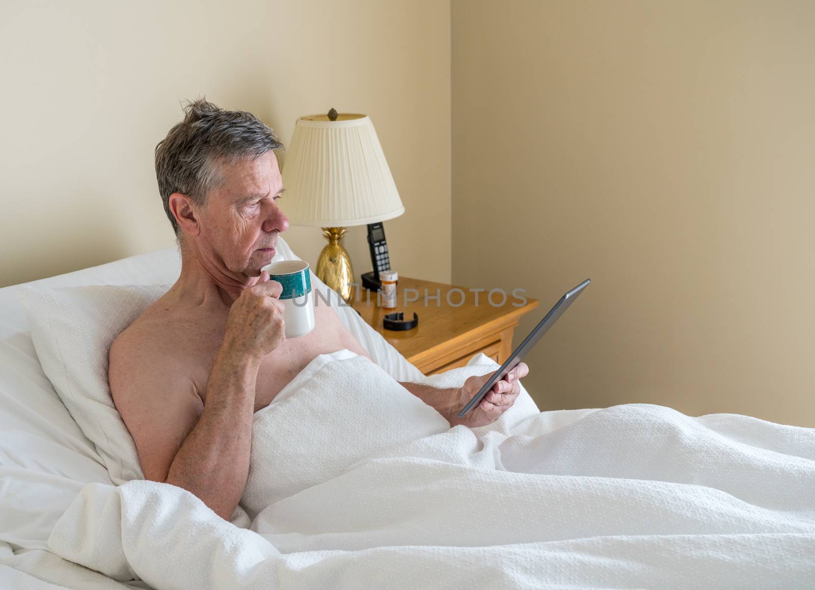 Senior retired caucasian man lying in adjustable bed on incline. He is reading and scrolling on digital tablet