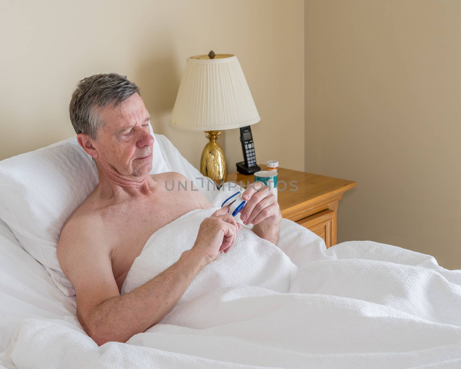 Senior retired caucasian man lying in adjustable bed on incline. He is checking his blood oxygen level with oximeter for fever or virus