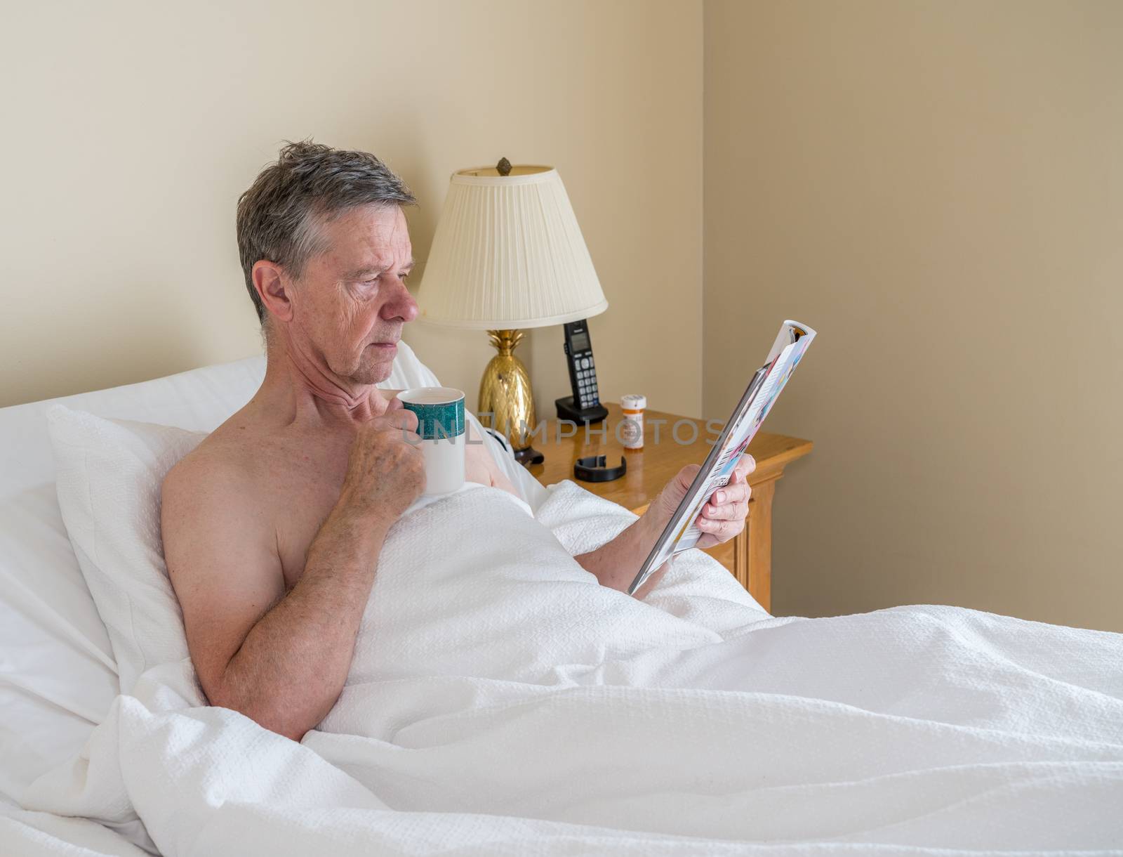 Single caucasian senior man in inclined bed and reading from magazine by steheap