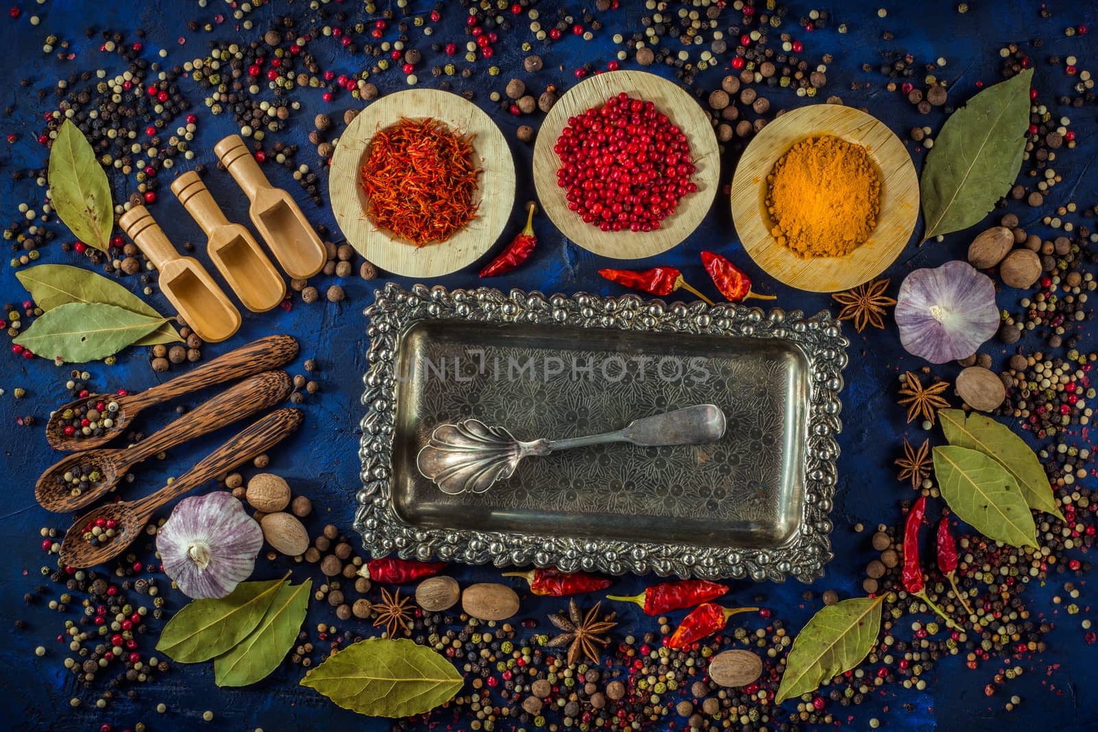 Assortment of colorful spices in the wooden bowls with spoons on a dark blue background. Curry, saffron, pink peppercorns, star anise, pepper, garlic, bay leaf and nutmeg with vintage silver plated tray with silver spoon. Top view. 