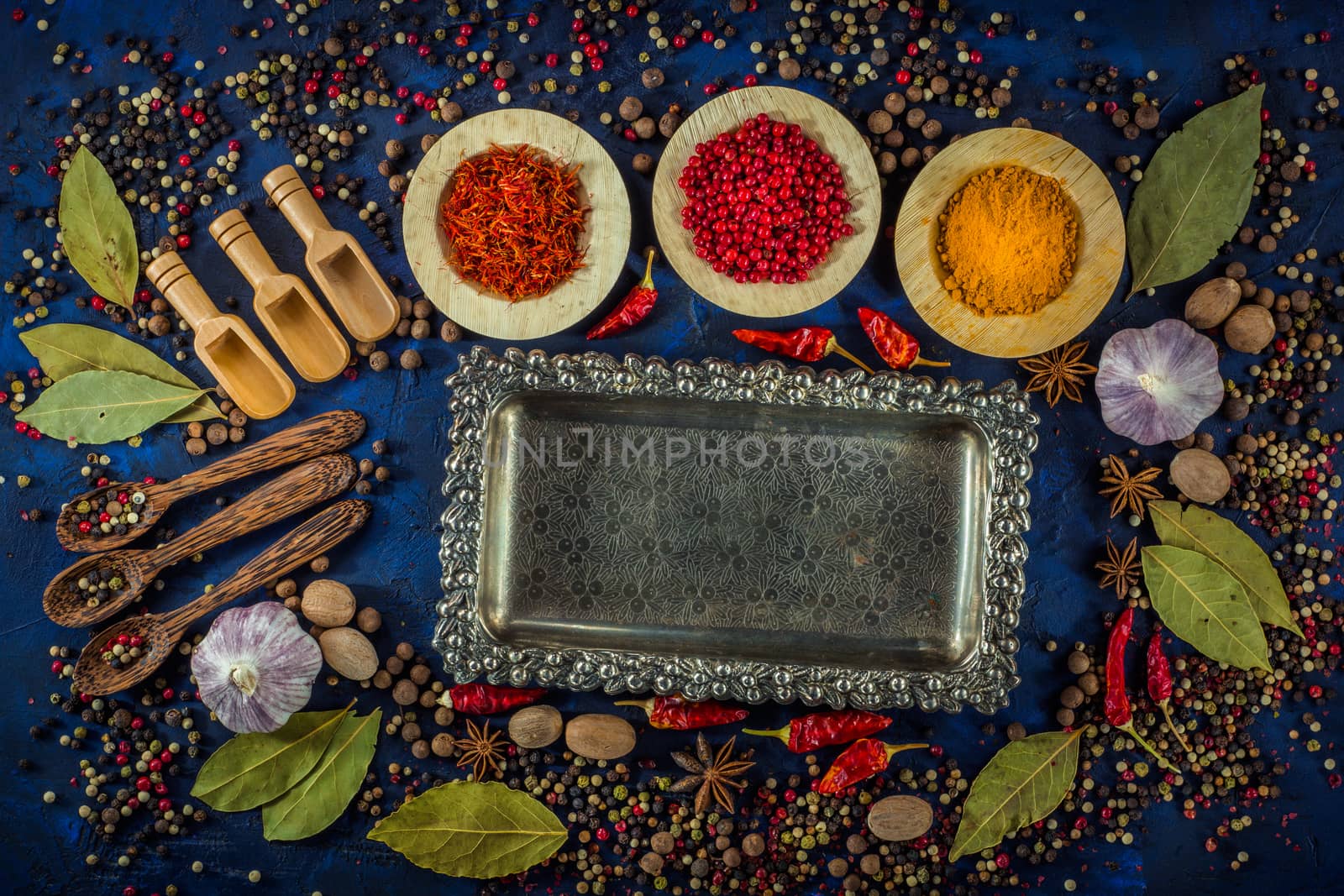 Assortment of colorful spices in the wooden bowls with spoons on a dark blue background. Curry, saffron, pink peppercorns, star anise, pepper, hot chili, garlic, bay leaf and nutmeg with vintage silver plated tray. Top view. 