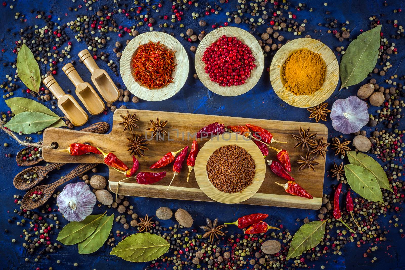 Assortment of colorful spices in the wooden bowls with spoons on a dark blue background. Curry, saffron, pink peppercorns, star anise, pepper, hot chili, garlic, bay leaf and nutmeg with cutting board. Top view. 