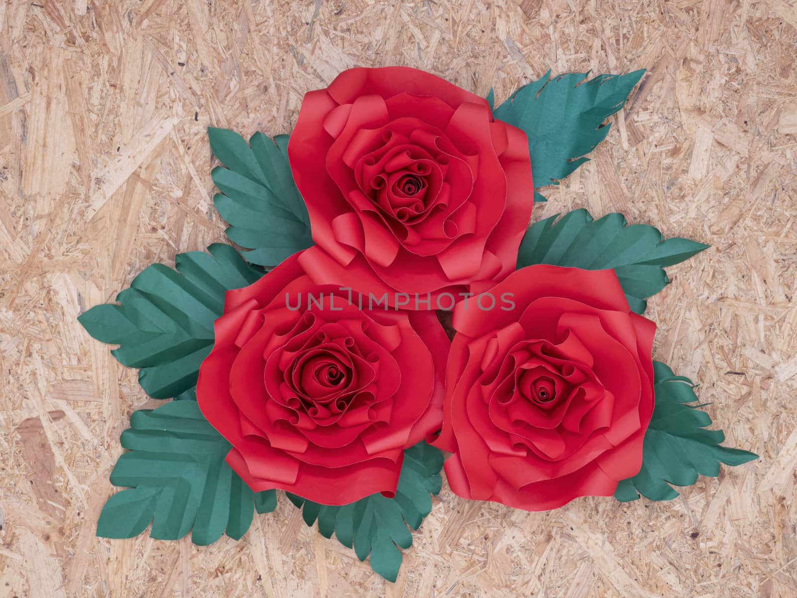 Beautiful hand-crafted paper crimson red roses with green leaves on vintage wooden board background.