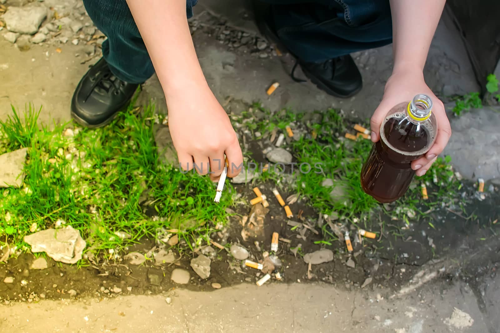 hands of a Smoking person who holds a bottle of alcohol, sitting on the street against the background of cigarette butts, garbage and trash scattered on the ground