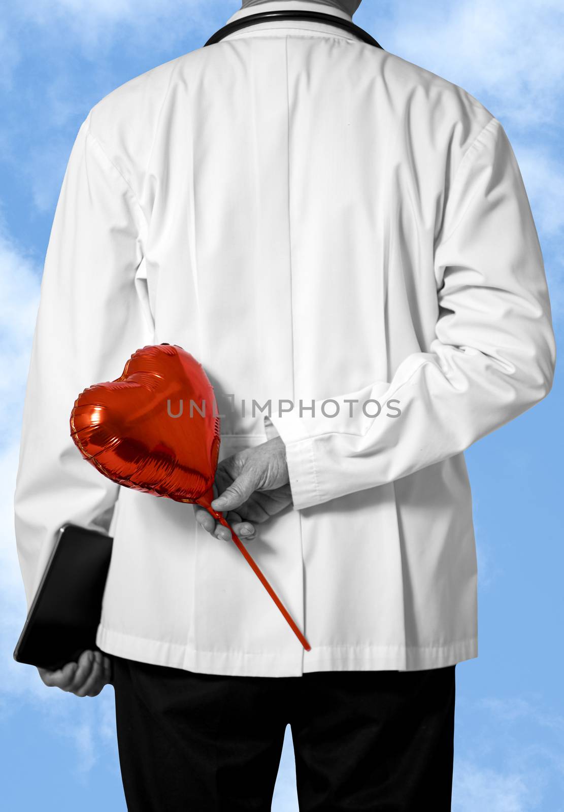 A male doctor in white coat holding a red balloon behind his back. Blue sky with white clouds background. Selective focus on red baloon with limited color. Doctor and caring concept.