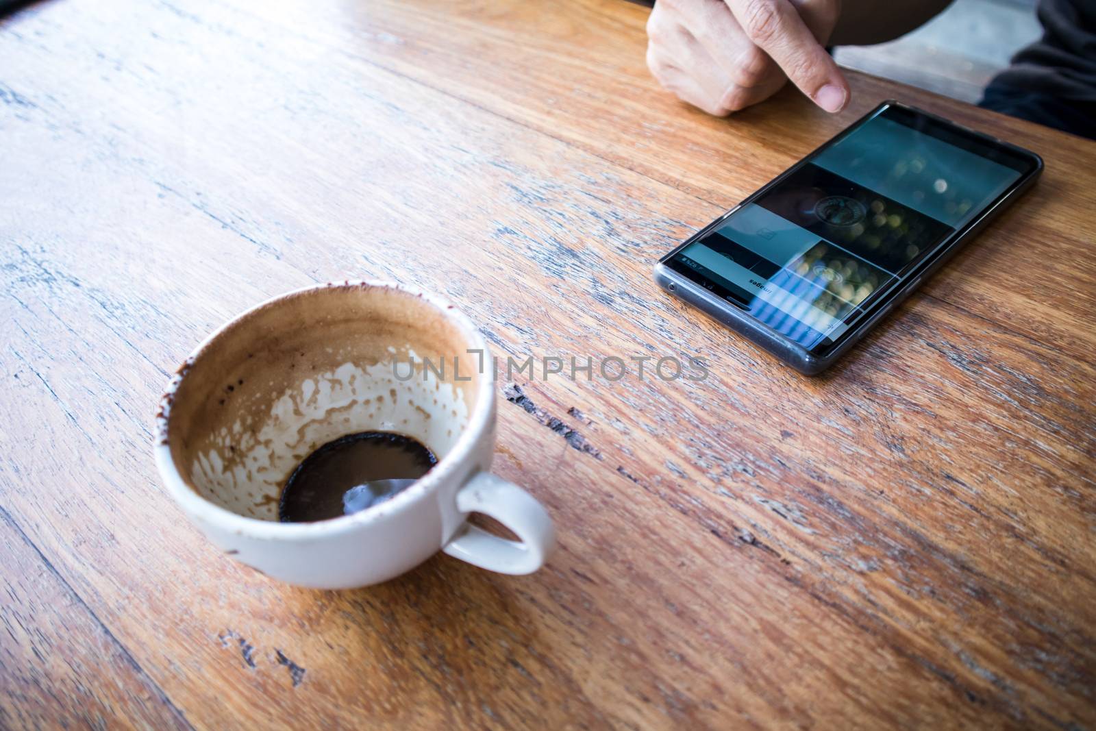 Close up of a right hand using smartphone with a Finished Empty Cup of Coffee on a brown wooden table. view from above