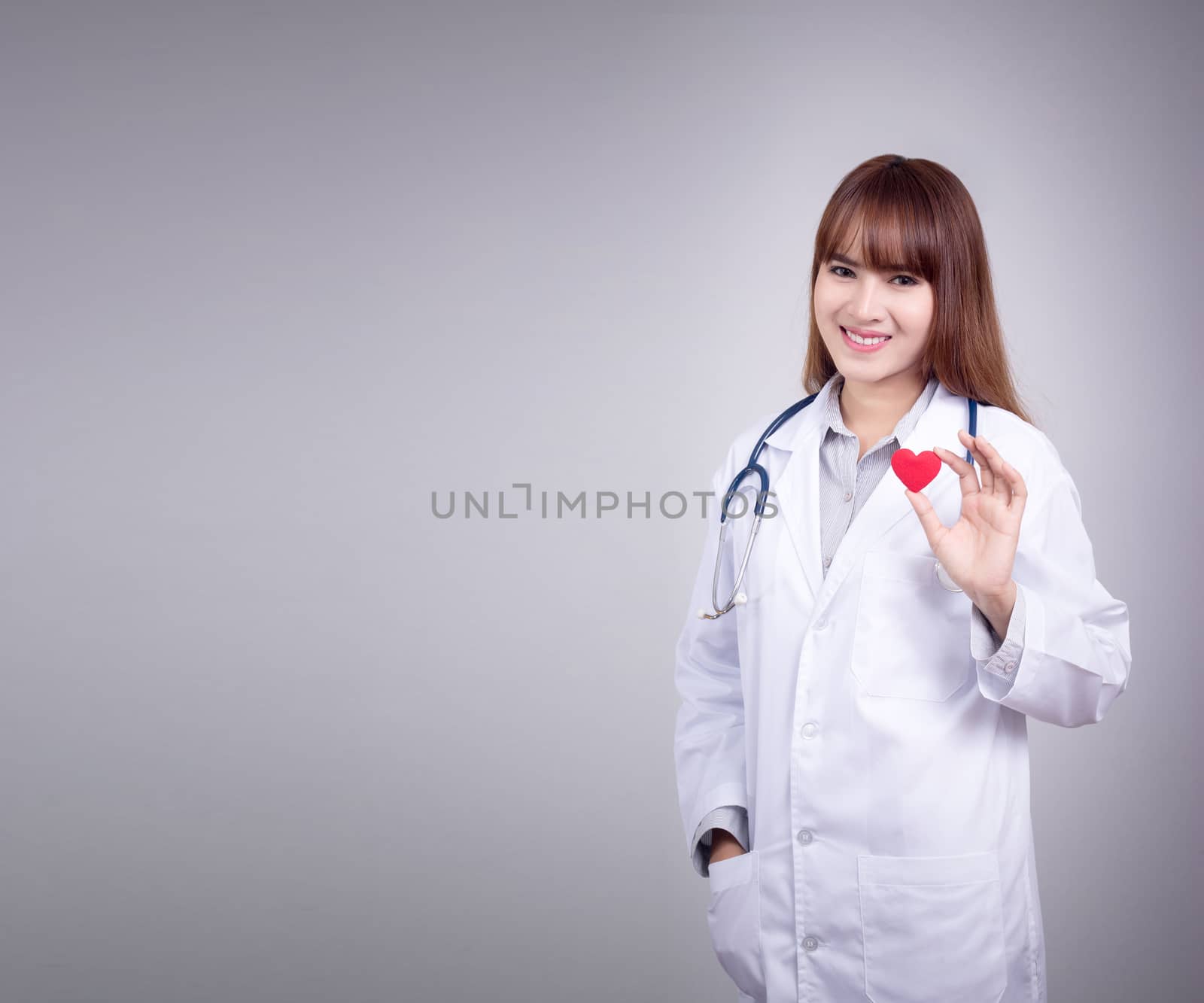 Young Asian doctor stand holding a red heart in her hand with smiley face. Dress up with doctor gown uniform with stethoscope in a hospital. Beautiful Asia female model in her 20s