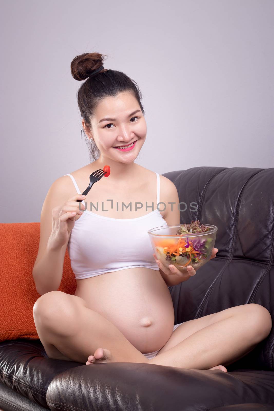 Pregnant nutrition healthy concept. Happy young beautiful Asian pregnant woman eat salad from salad bowl on sofa with smiley face. Health care with relax at home.Beautiful Asia female model in her 20s by asiandelight