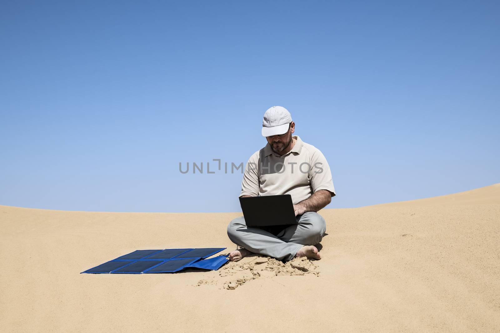 Adult (man) using his computer in the Middle of the dunes by clear sky, using portable flexible solar panels and charger