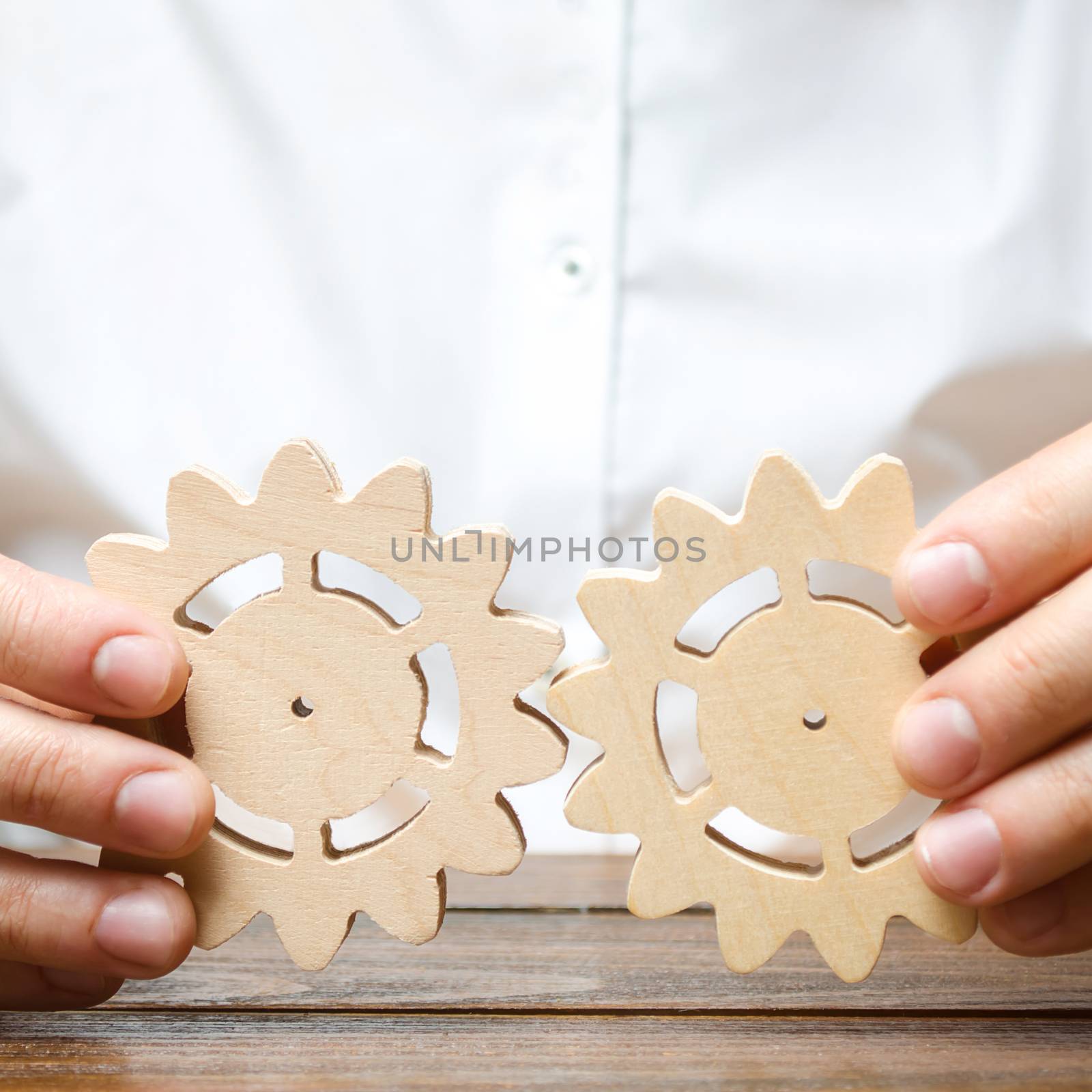 Businessman in white shirt connects two wooden gears. Improving work efficiency, establishing new connections and suppliers. Symbolism of establishing business processes and communication. by iLixe48