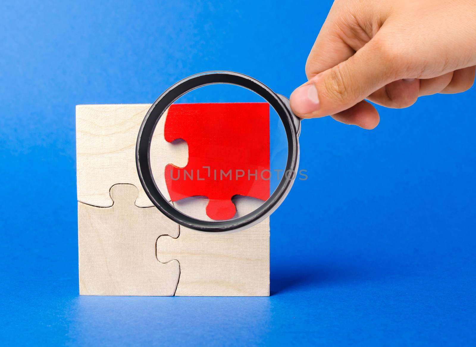 A magnifying glass looks at a red wooden puzzles on a blue background. Individual opinion. Stand out from the crowd. Uniqueness. Divergent views. Different concepts to other people. by iLixe48