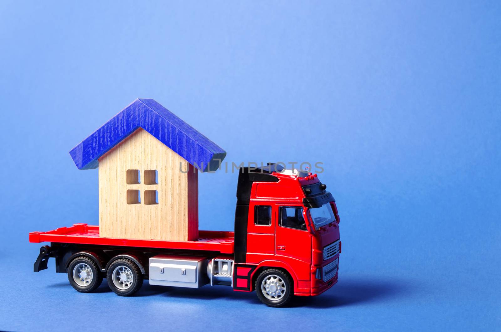 Red truck carrier transports a blue roofed house. Concept of transportation and cargo shipping, moving company. Construction of new houses and objects. Logistics and supply. Move entire buildings by iLixe48