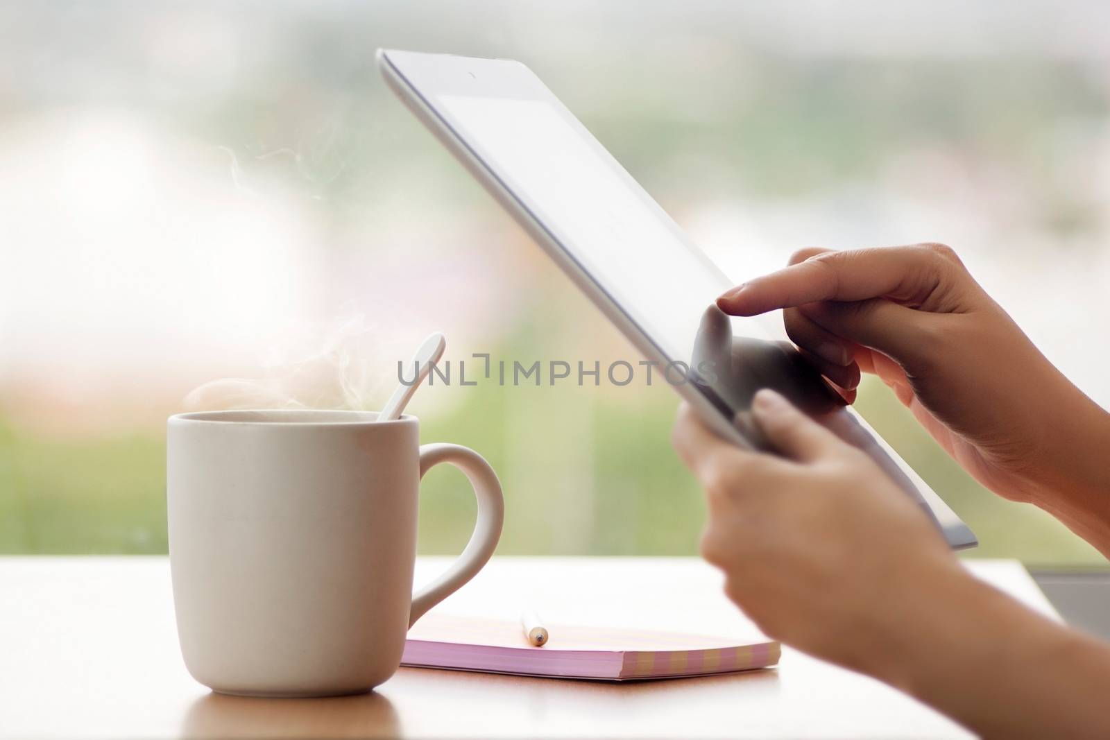 Tablet close up photo with woman hand. Girl using tablet in the cafe and drinking hot coffee. Woman using tablet computer laptop and make note on book with pencil with window glass background