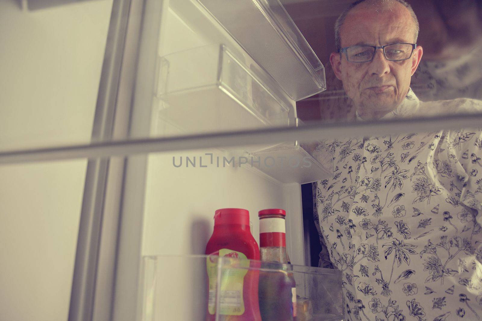 Senior man opening a refrigerator with little food. Selective focus. Hungry man looking for a late night meal in an empty fridge.