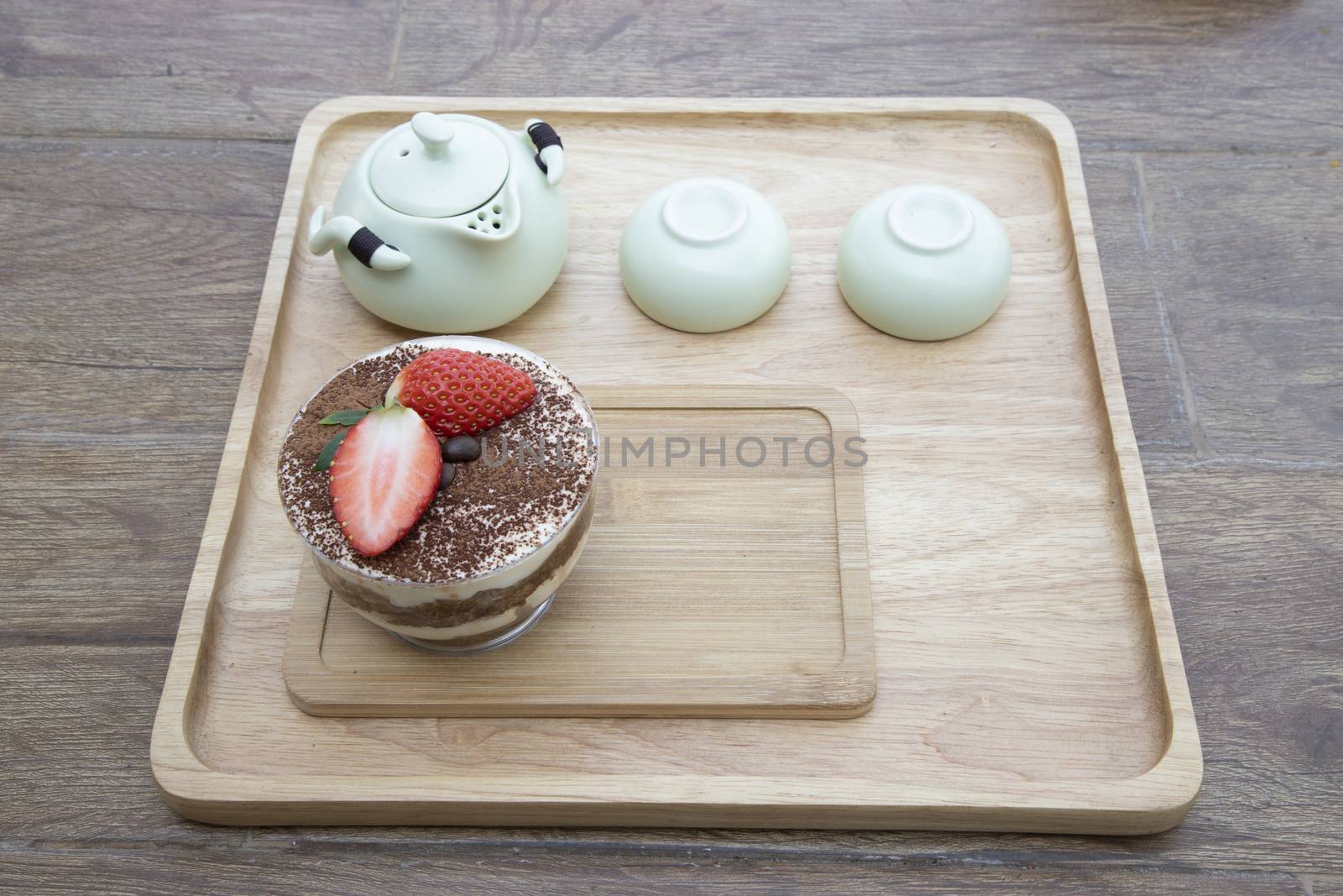 A cup of tiramisu cake, topped with fresh strawberry and coffee beans on a wooden tray with green ceramc tea pot set. Afternoon tea concept.