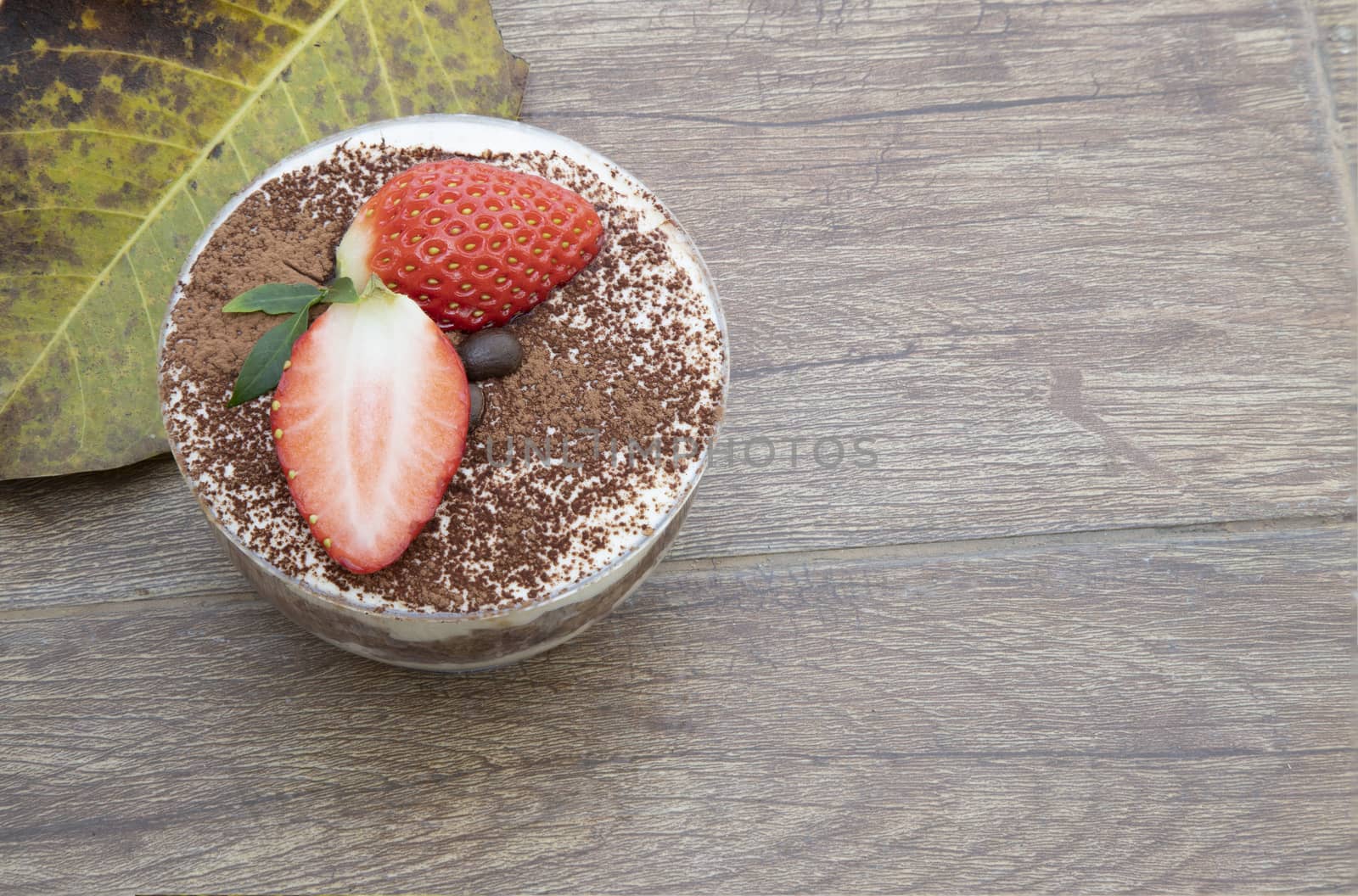 A cup of tiramisu cake, topped with fresh strawberry and coffee beans on a brown wooden table