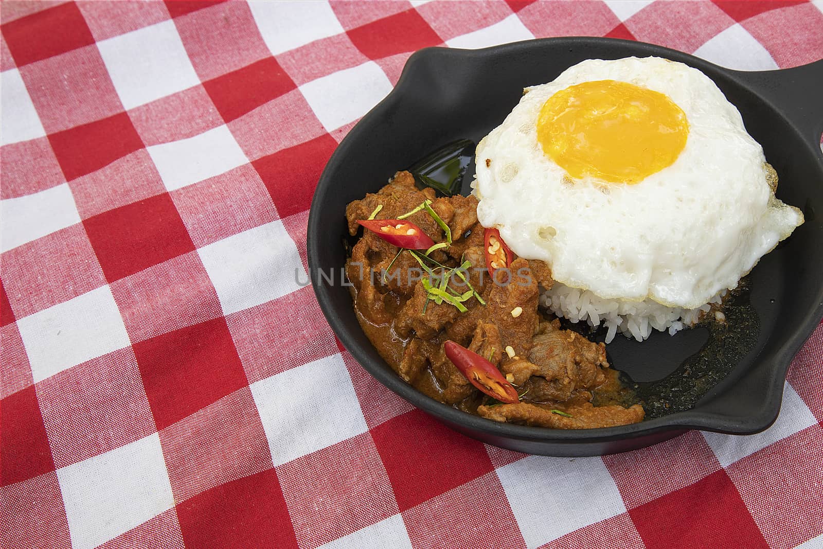 A serving of panang pork, Thai traditional red curry, with rice and fried egg in dark iron pan. Red and white checker pattern cotton cloth background.