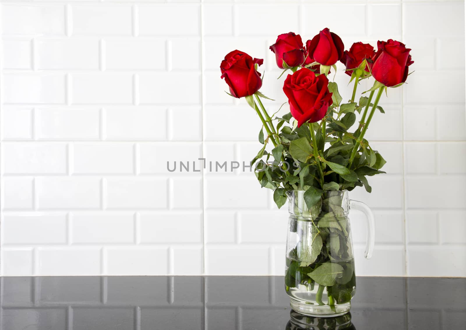 Beautiful red roses in a glass jug on black granite kitchen counter and white tile background.
