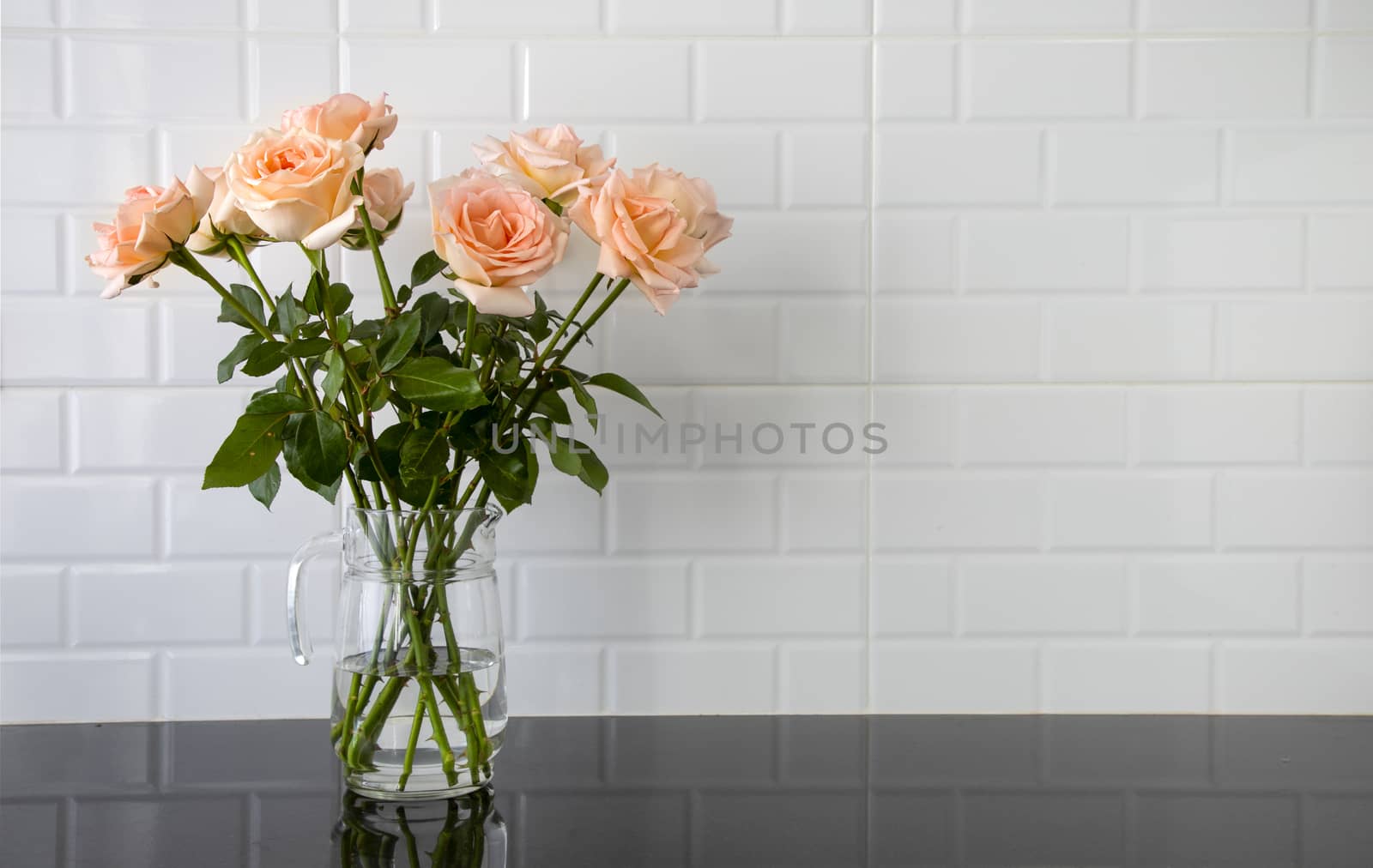 Beautiful peach color roses in a glass jug on black granite kitchen counter and white tile background.