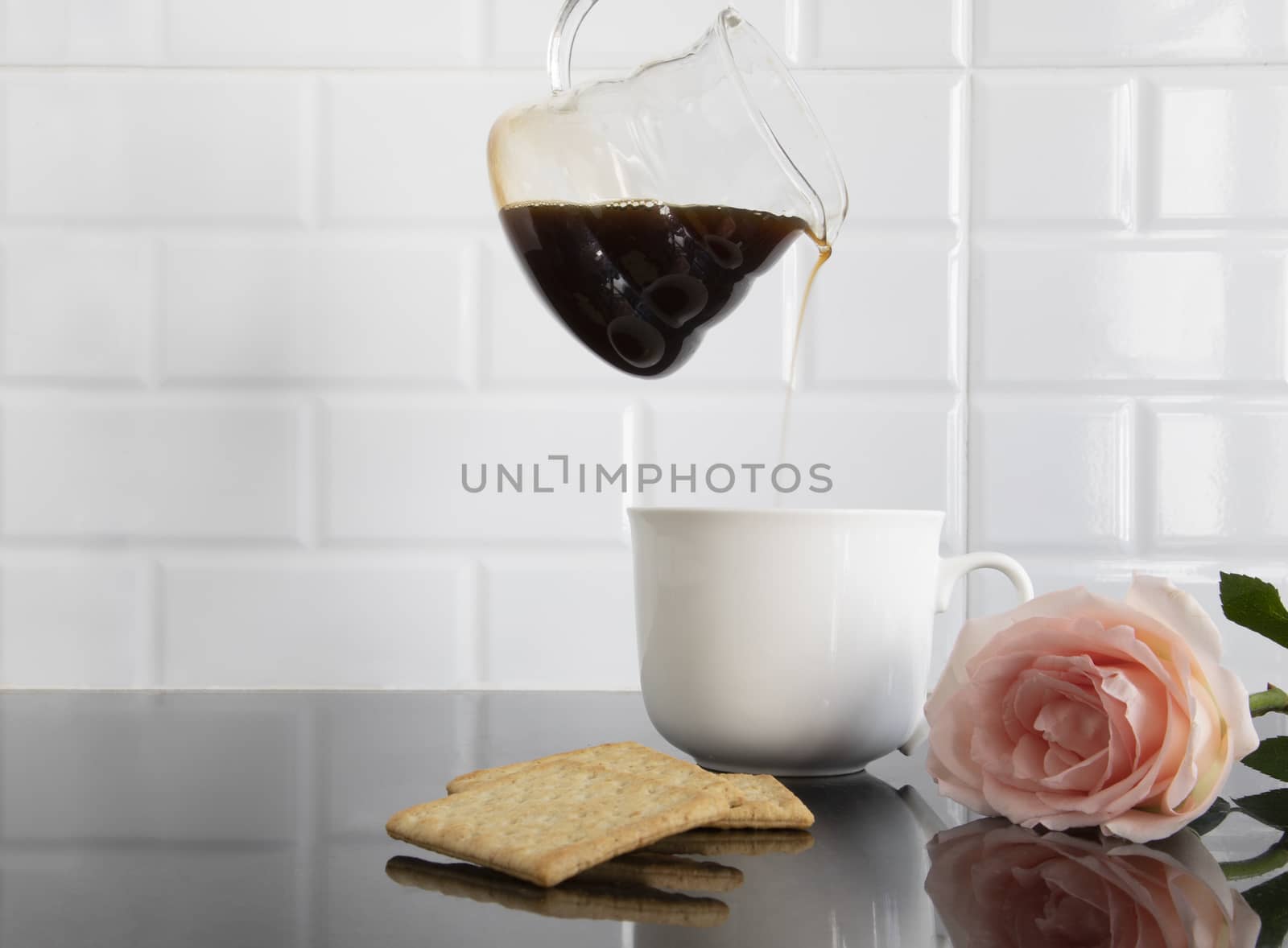 Image of someone pouring coffee to a white ceramic cup with pieces of biscuit and one peach color rose on a black granite kitchen counter.
