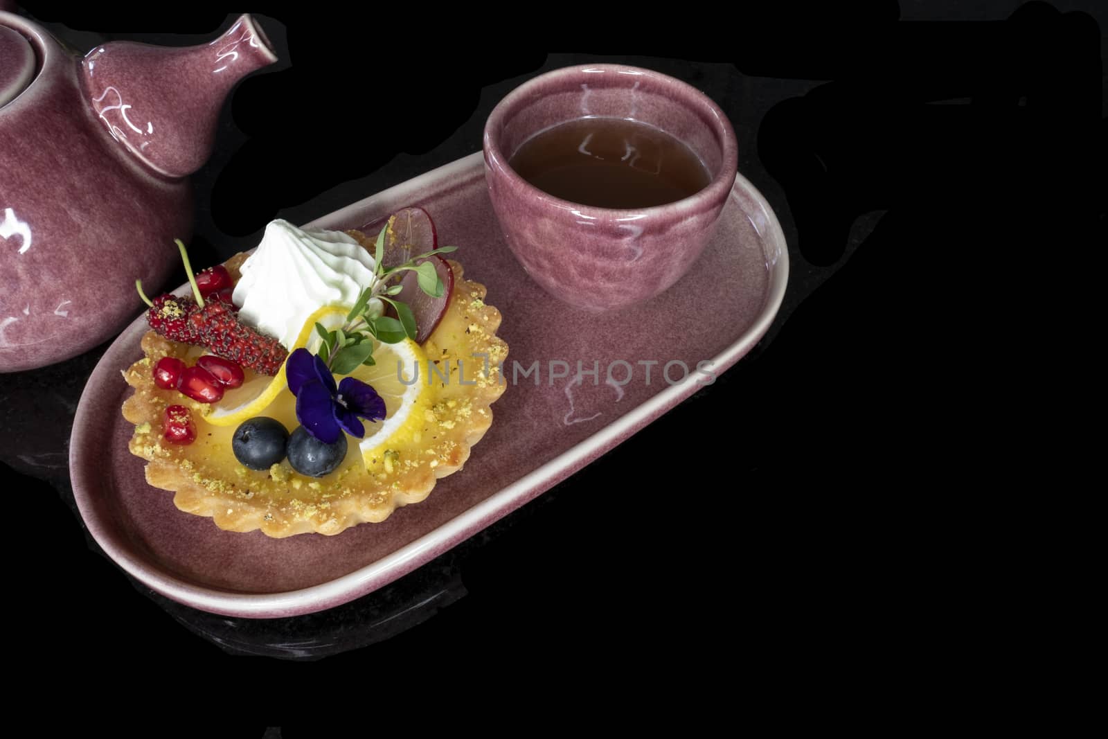 A piece of mixed fruit lemon tart on a purple ceramic plate with a tea cup and a chinese style ceramic tea pot. Black background.