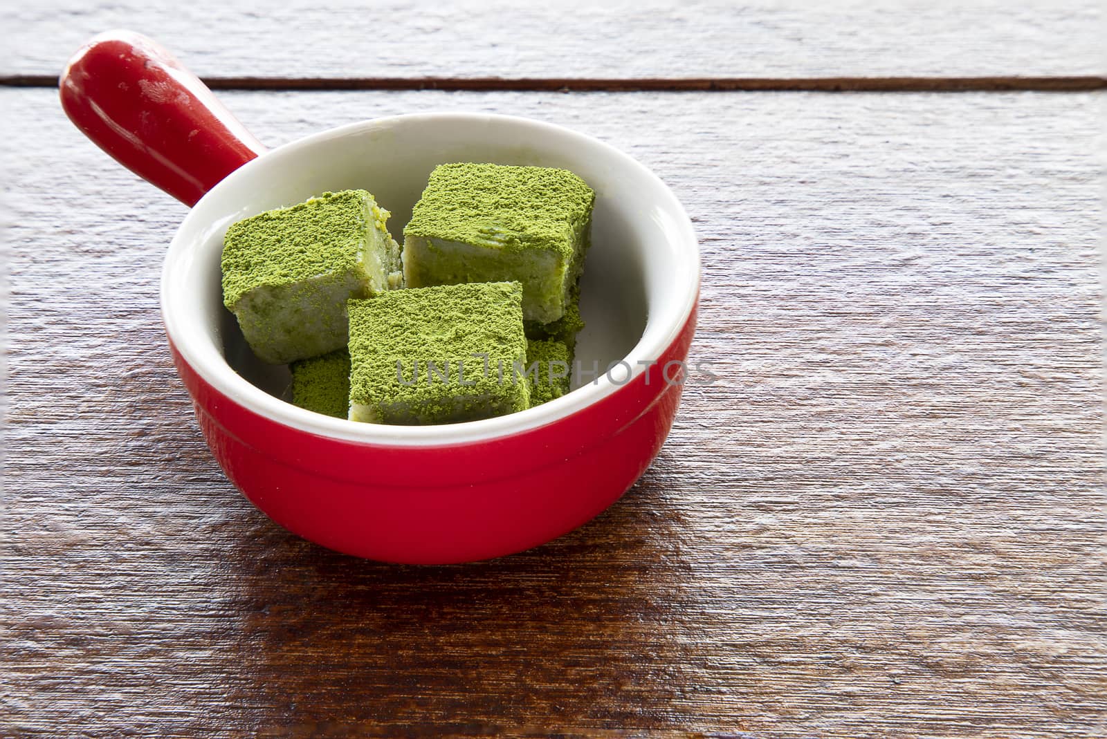 White chocolate fudge cubes heavily coated with fine green tea powder in a red ceramic dish. Brown wooden background.