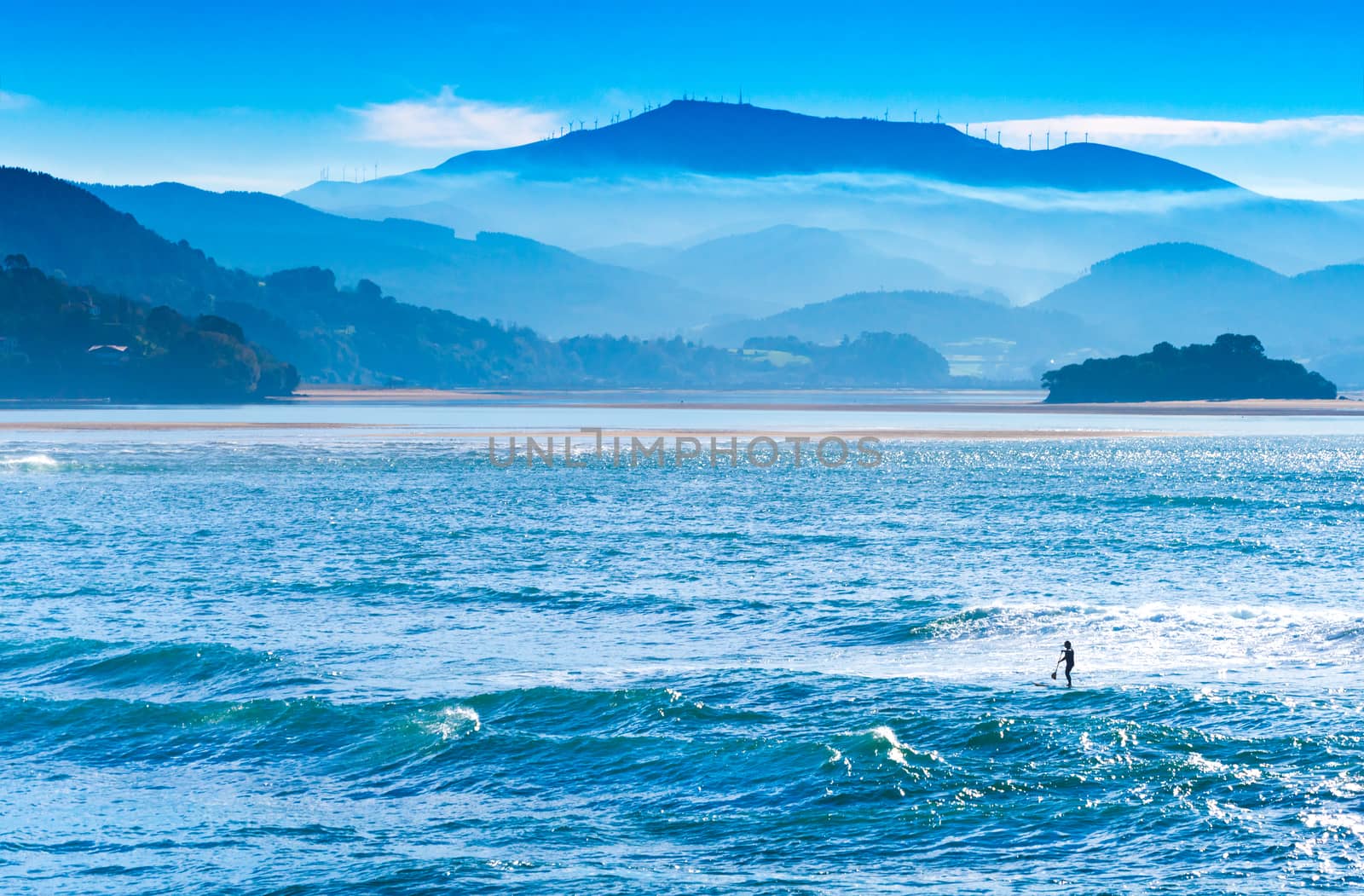 Beautiful landscape with foggy blue hills, ocean, and man on a surfboard