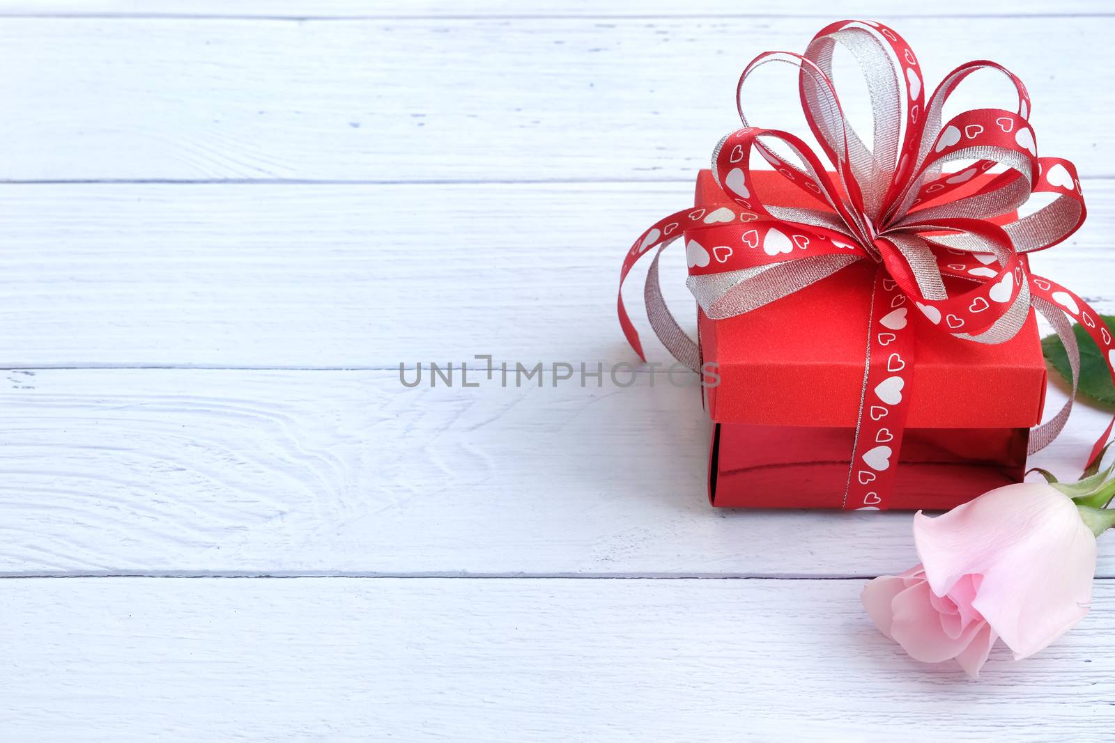 A red gift box with red and white ribbons printed with red and white hearts and a single blooming beautiful pink rose on a white wooden background. Love. Romance, Valentines' Day, Anniversary.