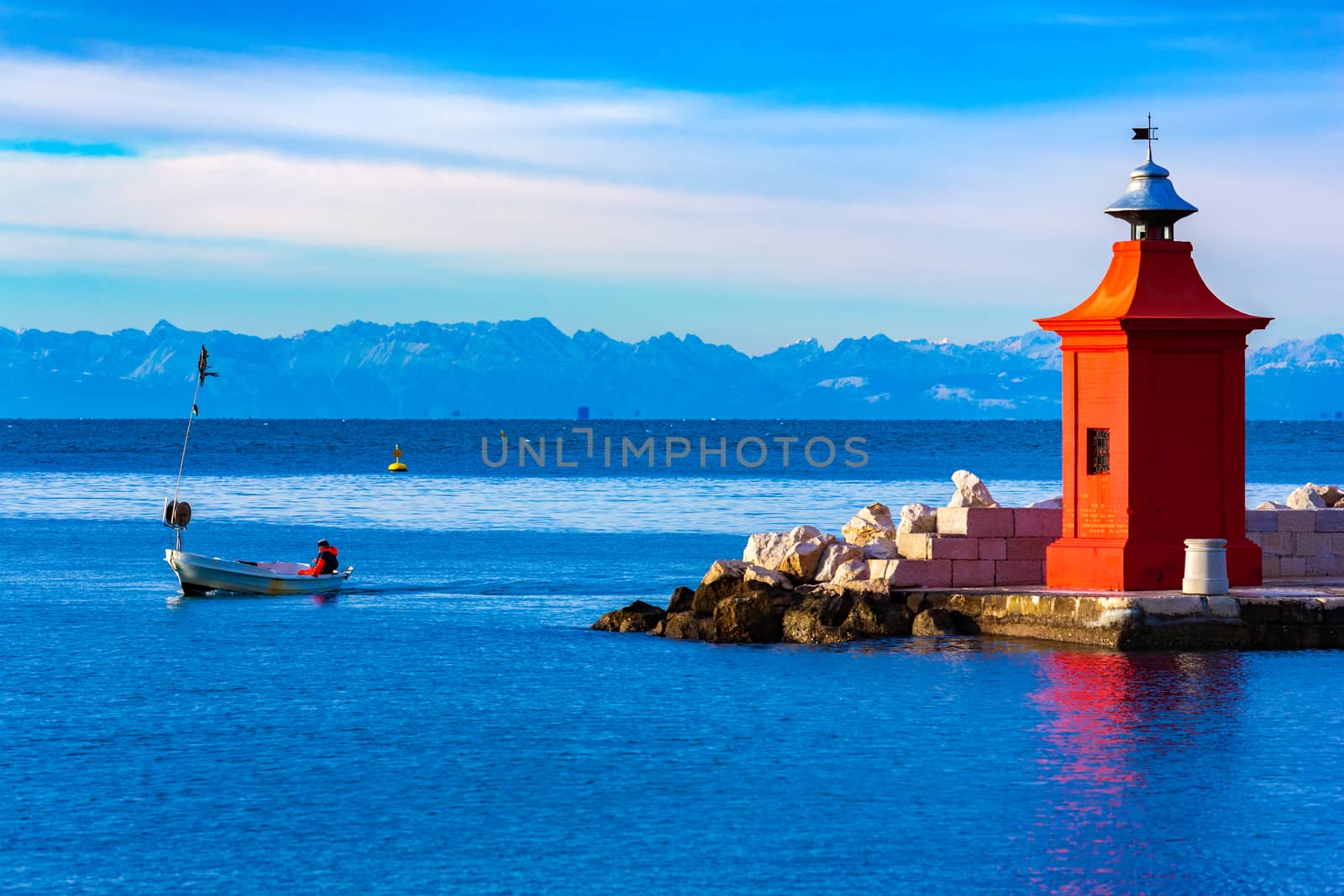 View of the red lighthouse on a pier, a fisherman on a boat and the beautiful mountain range on the horizon, Piran, Slovenia