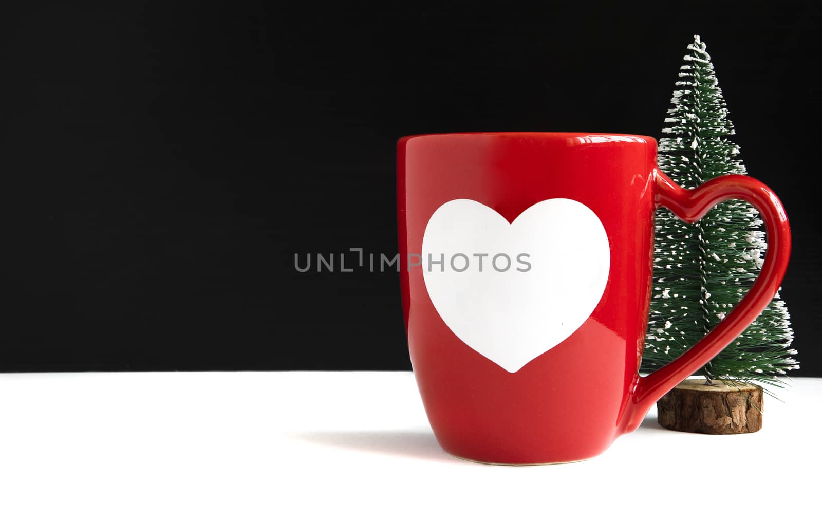 An orange ceramic mug with a printed white heart and a miniature Christmas tree on a table. Black and white background with copy space. Holidays, Christmas or New Year mockup.