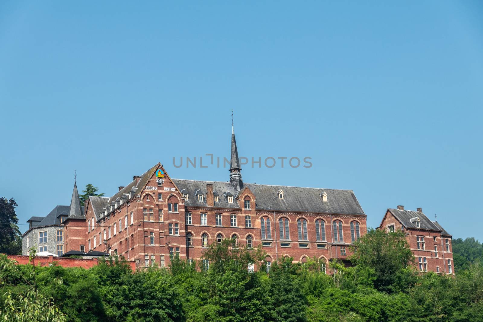 Dinant, Belgium - June 26, 2019: Red stone ancient convent of the Capuchins, now hotel, museum and administratif building under blue sky and green foliage in front.