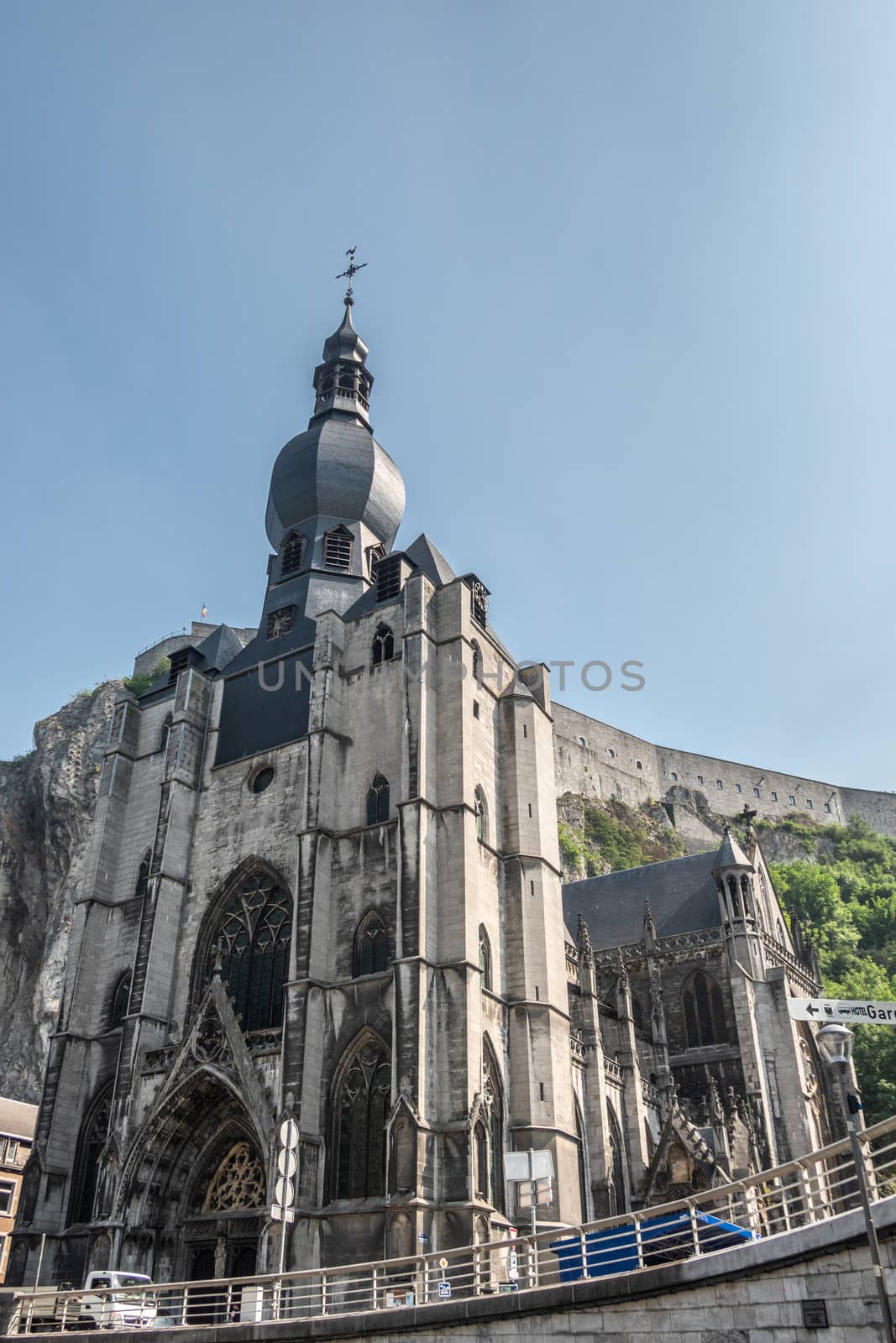Dinant, Belgium - June 26, 2019: The citadel fort on cliff view is parlty blocked by gray stone Notre Dame church under light blue sky. Fish eye view.