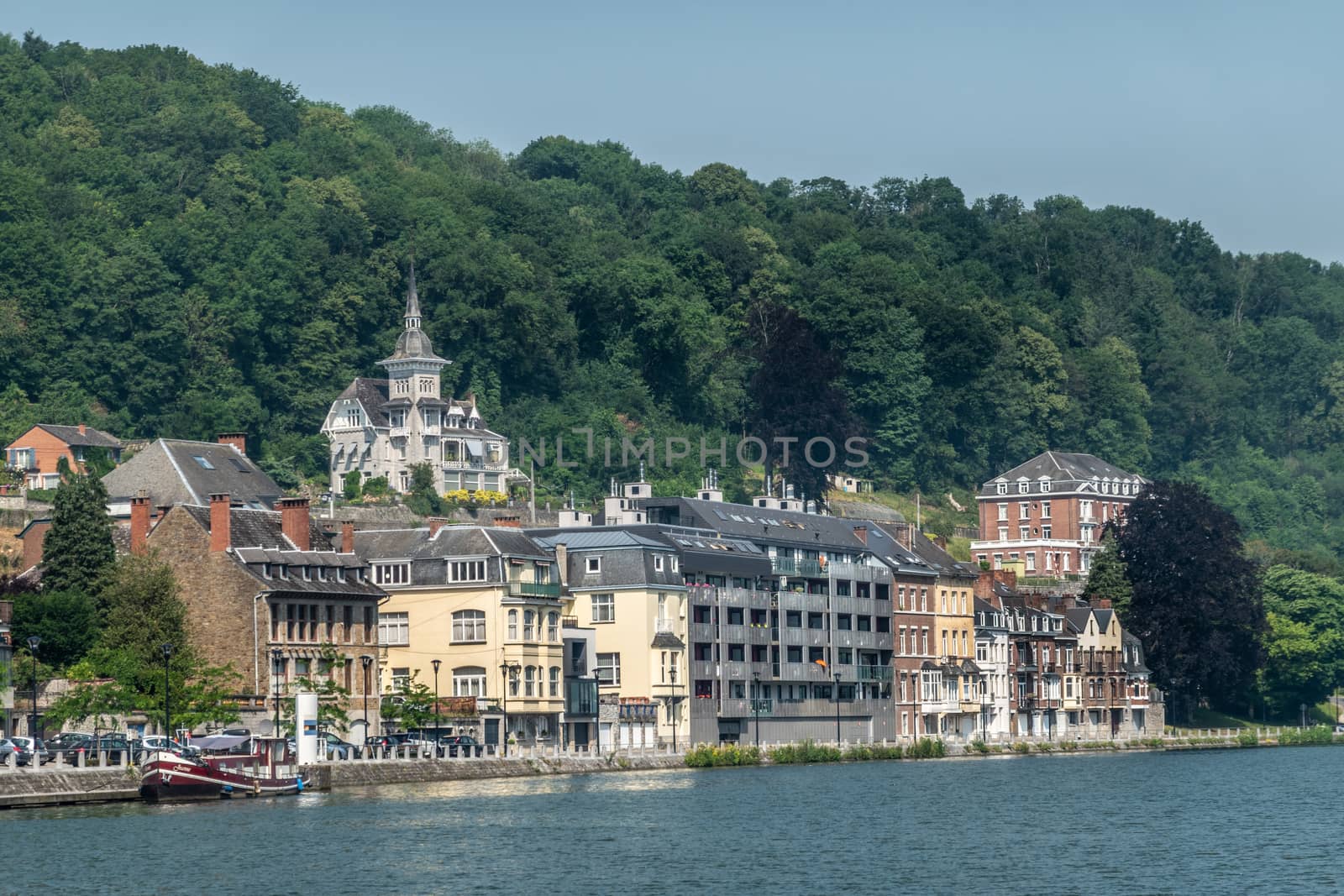 Dinant, Belgium - June 26, 2019: Left bank, north flow of Meuse River with buildings backed by green foliage and blue sky. Tower on mansion in forest foliage.