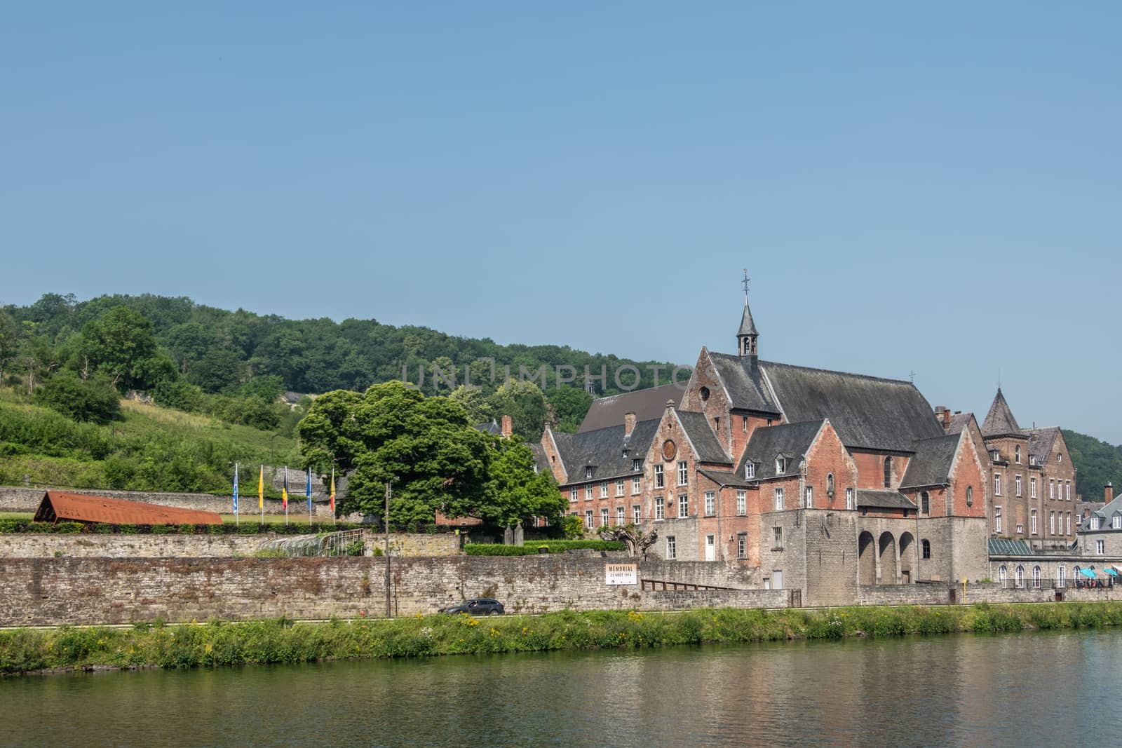 Dinant, Belgium - June 26, 2019: Part of Red stone ancient convent of the Capuchins is now the seat of CPAS, the social services of the city. Meuse River in front, blue sky.