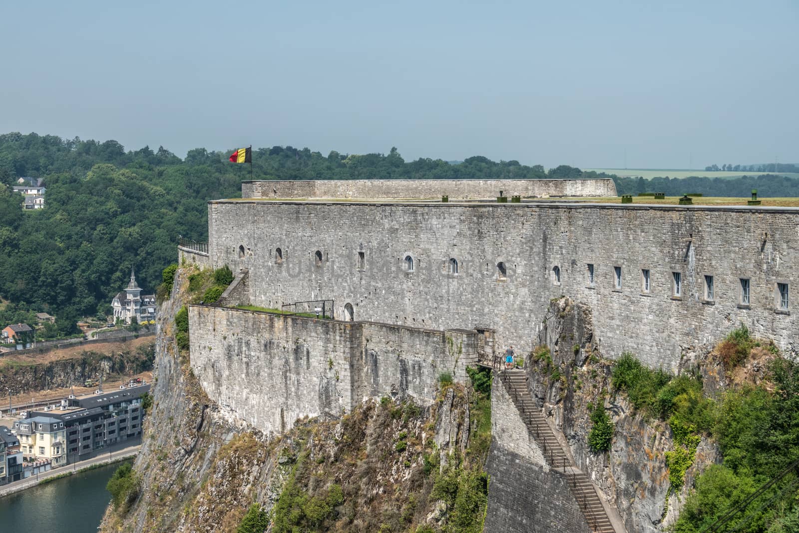 Dinant, Belgium - June 26, 2019: Southern gray stone ramparts of Citadelle above the city with Belgian flag under blue sky. Green forests in back. Meuse River down.