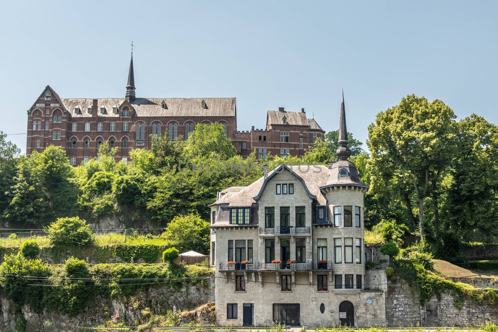 Dinant, Belgium - June 26, 2019: Red stone ancient convent of the Capuchins, now hotel, museum and administratif building and Villa Mouchenne, top restaurant, in front. Blue sky, green foliage.