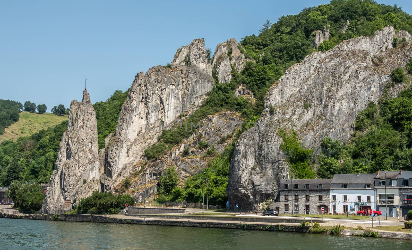 Dinant, Belgium - June 26, 2019: Wider view on South side of Le rocher Bayard along La Meuse River surrounded by green foliage under blue sky.