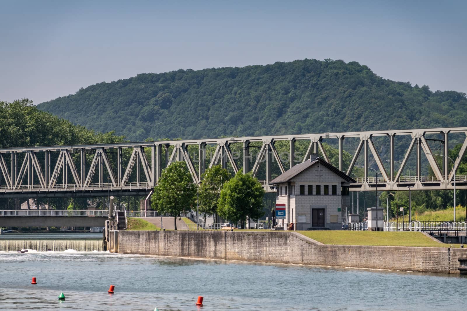 Dinant, Belgium - June 26, 2019: Gray lock operator office building with train bridge span over it against forested hill and blue sky. Rapids to the left.
