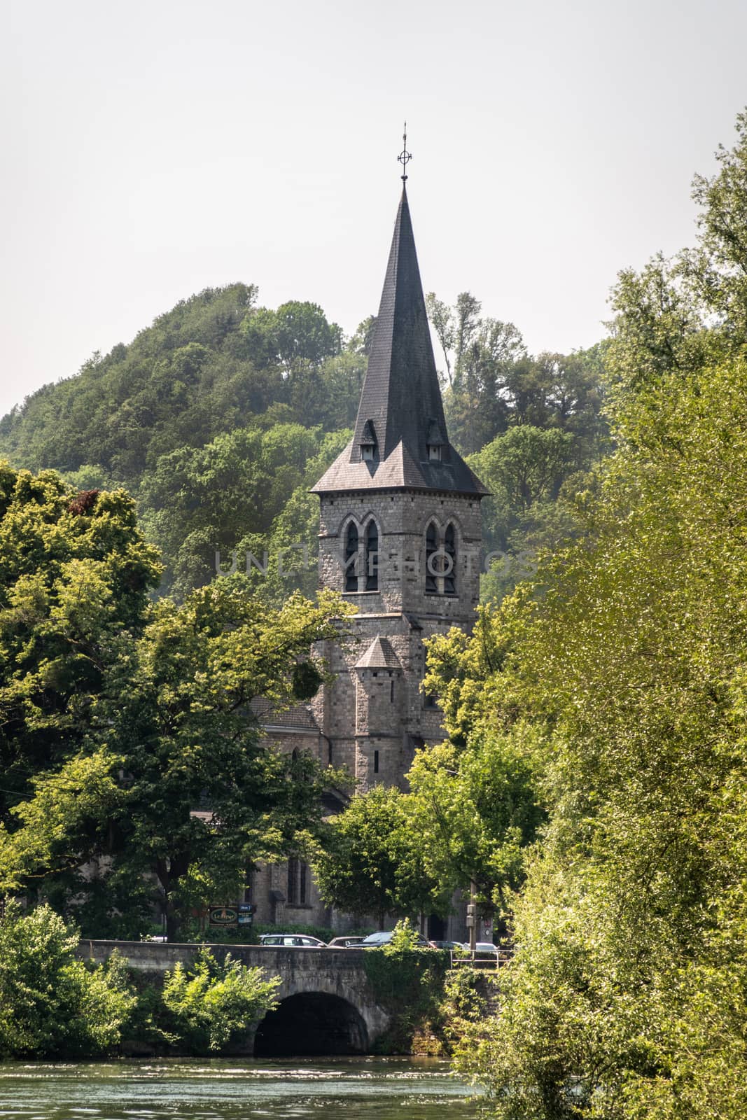 Dinant, Belgium - June 26, 2019: Gray stone tower and spire of Saint Anne church surrounded by trees and green foliage under silver sky. Old roman bridge over Lesse River.