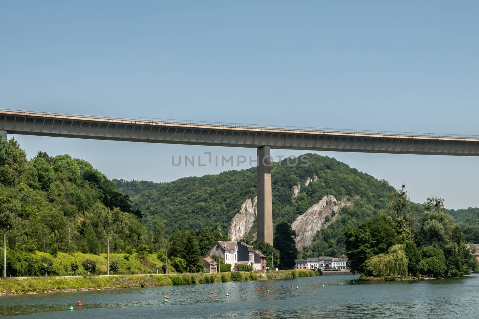 Dinant, Belgium - June 26, 2019: Very high Route Charlemagne, N97, concrete bridge over Meuse River under light blue sky with green forested hills and Ile d’ Amour in center of river.