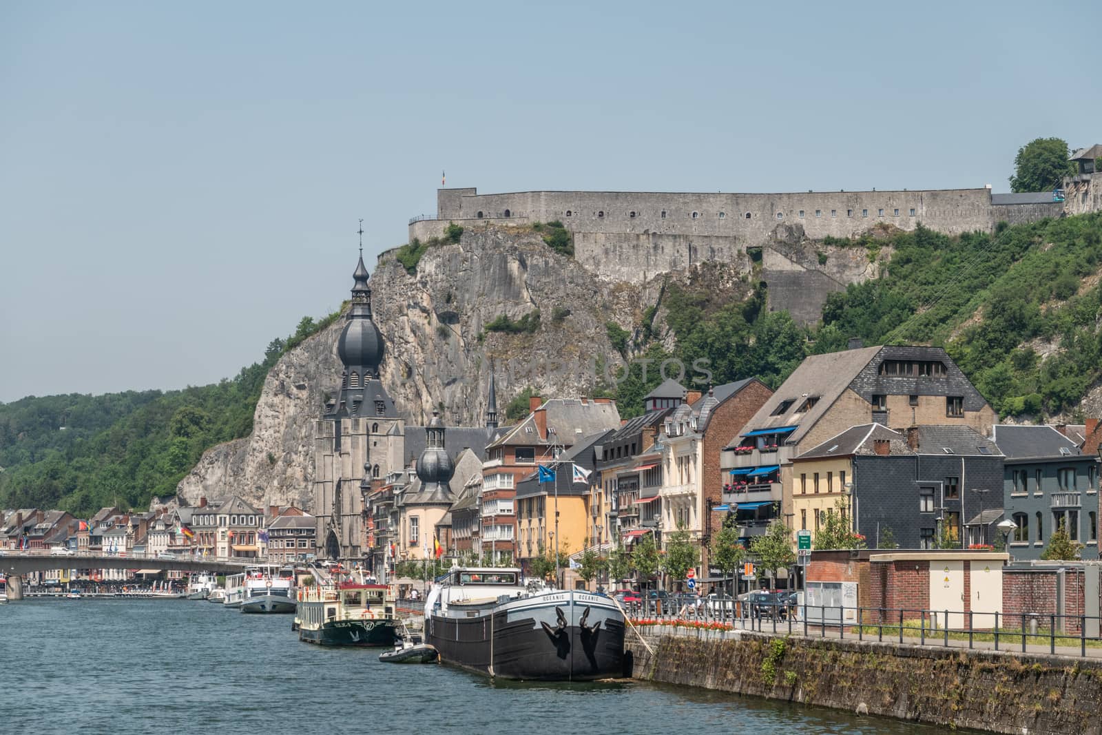 Dinant, Belgium - June 26, 2019: Citadelle fortification seen from right south bank of Meuse River with church and buildings, cars, boats under blue sky.
