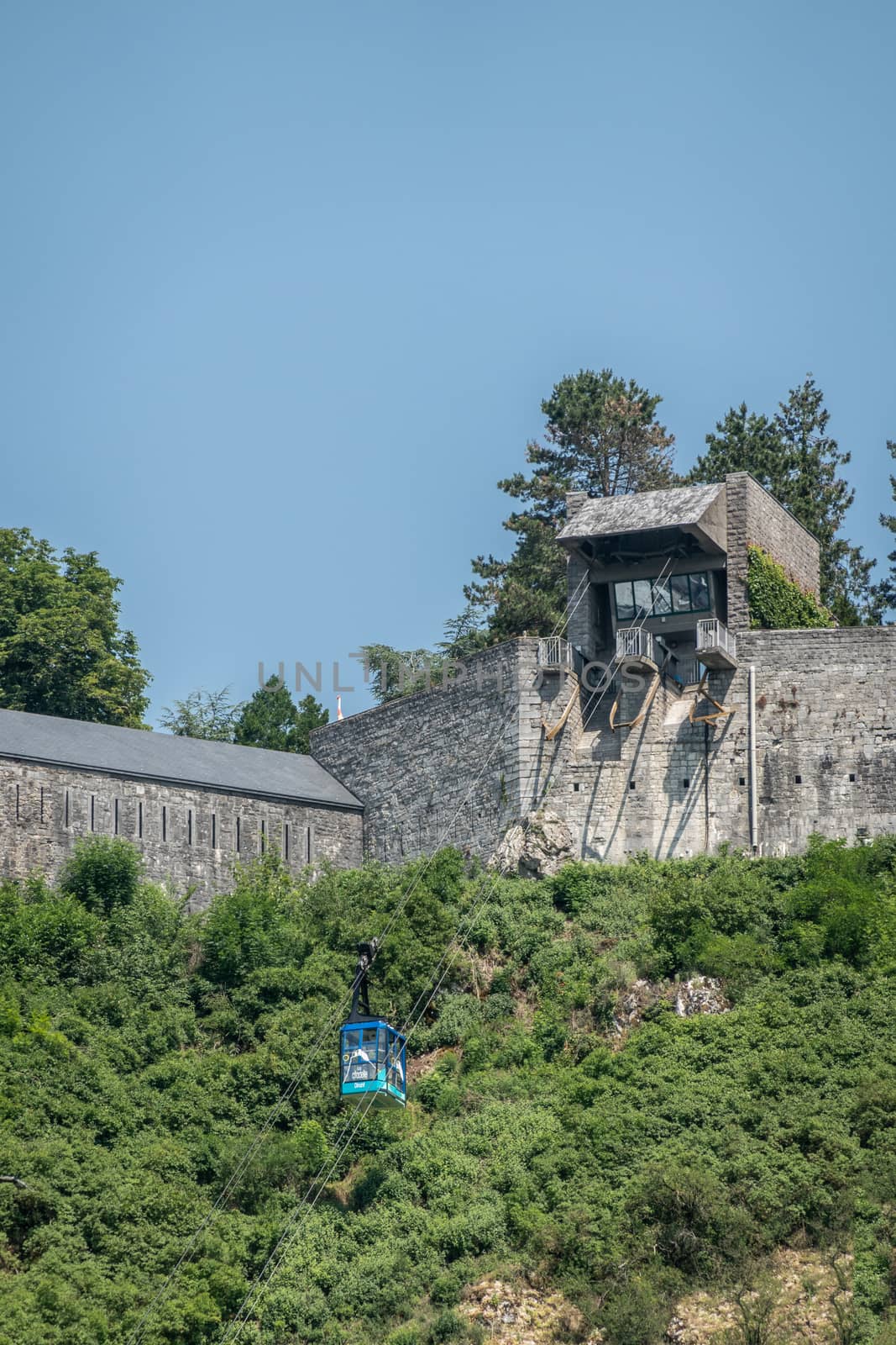 Dinant, Belgium - June 26, 2019: Blue cable car against green foliage bringing visitors up along gray stone cliffs and ramparts of Citadelle under blue sky.