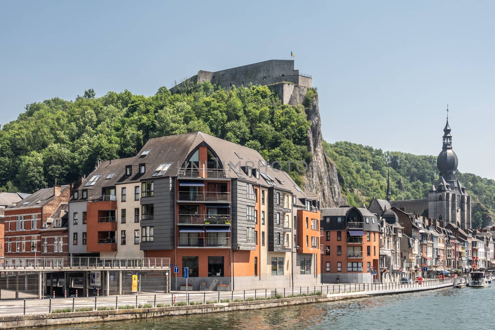 Dinant, Belgium - June 26, 2019: Citadelle fortification seen from right north bank of Meuse River with church and buildings, cars, boats under blue sky. Green foliage.