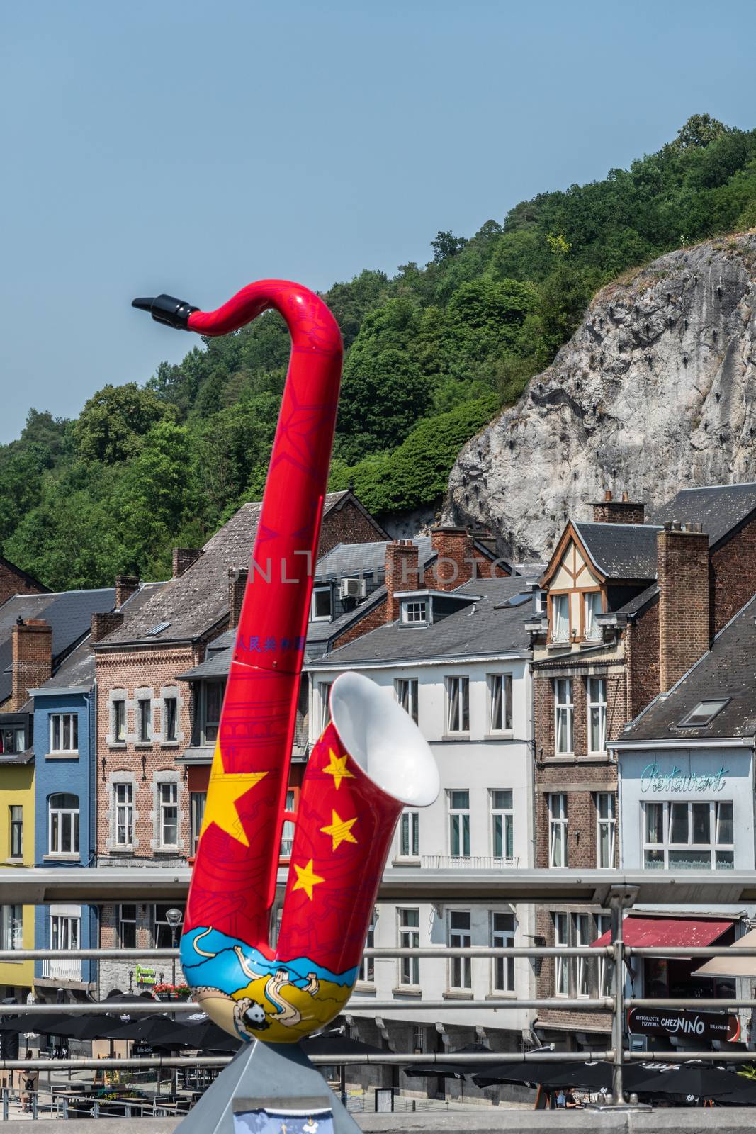 Dinant, Belgium - June 26, 2019: Red and Yellow saxophone statue to honor China on Charles de Gaulle bridge. Business and houses in back under green foliage and blue sky.